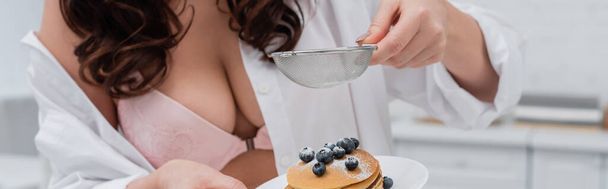 Cropped view of sexy woman in bra pouring powdered sugar on pancakes in kitchen, banner  - Photo, Image