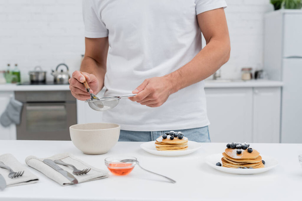 Cropped view of man pouring powdered sugar in sieve near honey and pancakes in kitchen  - Photo, Image
