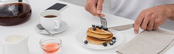 Cropped view of man cutting pancakes with blueberries near coffee and smartphone, banner  - Foto, Imagem