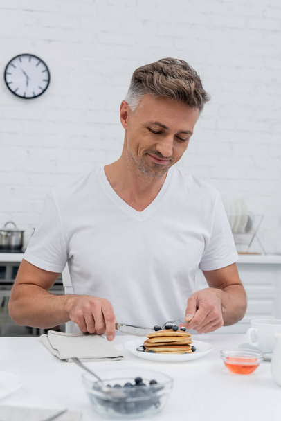 Positive man cutting pancakes near blurred blueberries and honey in kitchen  - Valokuva, kuva