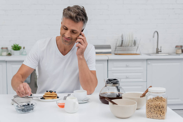 Smiling man talking on smartphone near delicious breakfast in kitchen  - Foto, Imagen