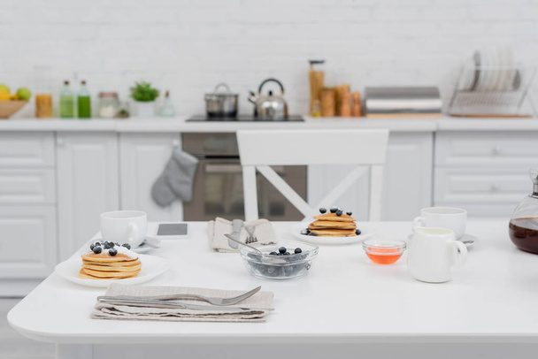 Tasty pancakes with blueberries near coffee and smartphone in kitchen  - Photo, Image