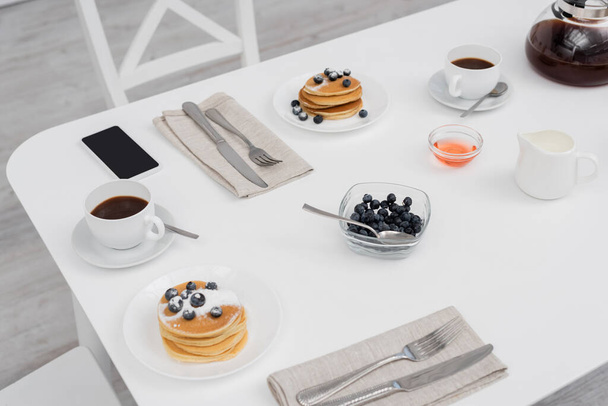 Blick auf das Smartphone in der Nähe von Kaffee und Pfannkuchen in der Küche  - Foto, Bild