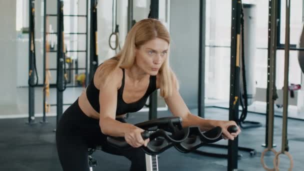 Mujer atleta motivada de mediana edad que hace ejercicio en el aparato de entrenamiento de bicicleta en el gimnasio, preparándose para la maratón de bicicletas, tiro de cerca, espacio libre - Imágenes, Vídeo