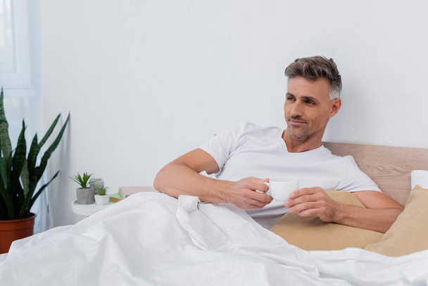 Man in t-shirt holding cup of coffee while lying on bed at home  - Fotoğraf, Görsel