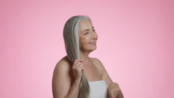 Speciale haarverzorging. Mooie senior dame verpakt in handdoek borstelen haar grijze haar met speciale kam na het schoonmaken, genieten van styling, roze studio achtergrond, slow motion - Video