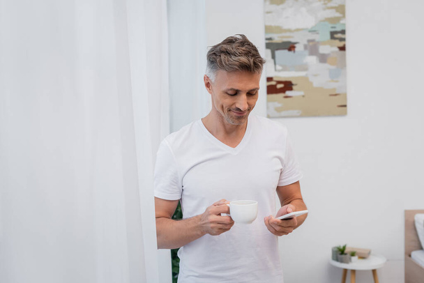 Positive man in t-shirt holding cup of coffee and using smartphone at home  - Foto, Bild