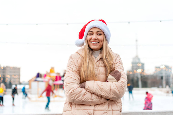 Outdoor portrait young beautiful fashionable woman wearing Santa hat, posing street of European city. Winter Christmas holidays concept. ice skate rink background. Caucasian female winter clothes - Fotó, kép