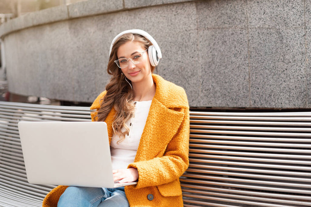 Business Woman Wear Eyeglasses Listen Music Headphone Outdoor Sitting Bench Using Laptop Outdoor Dressed Stylish Yellow Coat Smile Caucasian Female 30s Enjoy Podcast Or Audio Books Outside  - Fotoğraf, Görsel