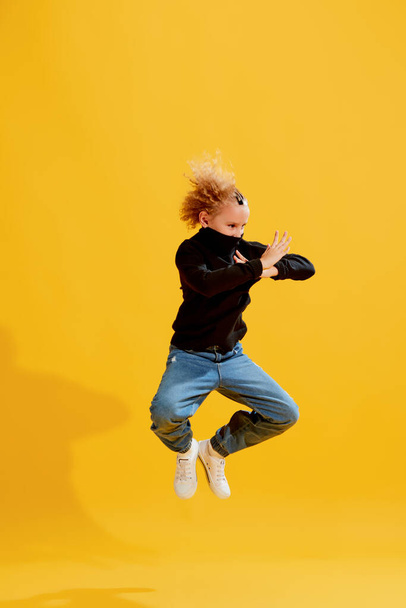 Expressive little girl, kid in black long sleeve blouse and jeans jumping isolated over yellow background. Kids emotions, news, challenges, childhood, education concept - Foto, Imagem