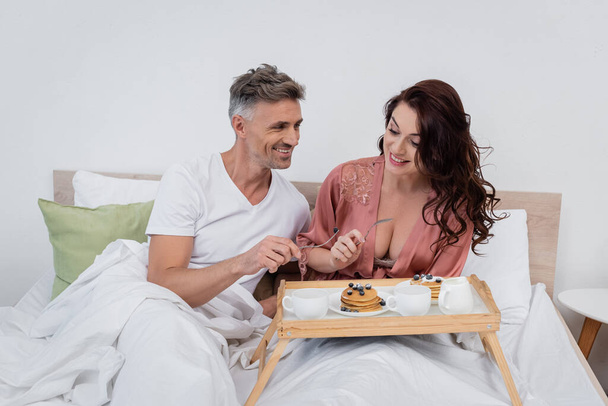 ベッドの上で朝食トレイにパンケーキやコーヒーの近くにフォークを保持笑顔カップル  - 写真・画像