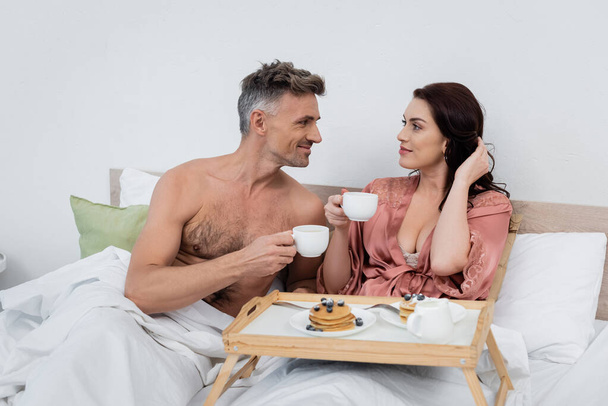 Hemdloser Mann hält Tasse Kaffee in der Nähe Frau in Seidenmantel und Frühstück auf Tablett im Bett  - Foto, Bild