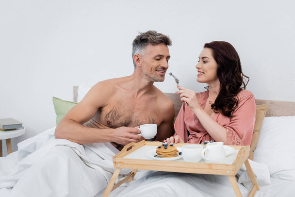 Cheerful woman in silk robe feeding husband with blueberry near breakfast on tray on bed  - Fotografie, Obrázek