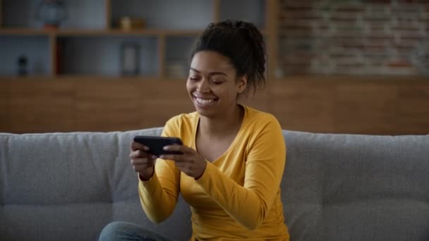 Concept de jeu en ligne. Jeune femme afro-américaine heureuse jouant à des jeux vidéo sur smartphone, profitant de cyberdivertissement à la maison, au ralenti, espace libre - Séquence, vidéo