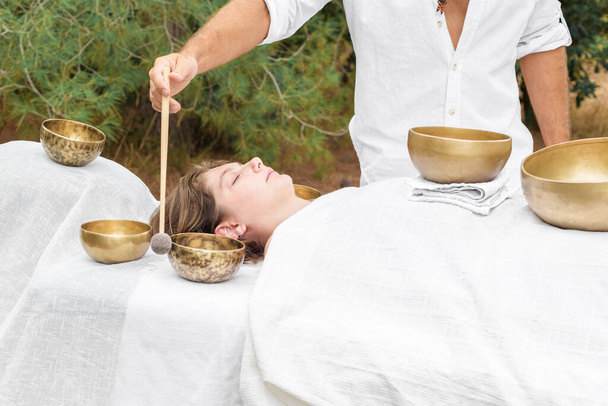 Young woman laying on a massage bed with tibetan singing bowl outdoor. A master of sound massage therapy doing a Buddhist healing practices, male hand holding a felt clapper - Photo, Image