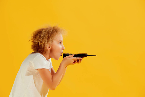 Voice message. Cute little girl, kid using retro mobile phone isolated over yellow background. Concept of children emotions, fashion, beauty, eras comparison. Copy space for ad - Photo, Image