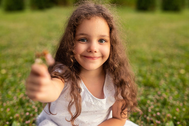 Adorable little girl in white dress outstretching hand with flower at camera sitting on grass in summer park - Photo, Image