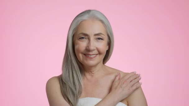 Coccole profonde del corpo. Studio ritratto di donna anziana positiva avvolto in asciugamano sfregamento crema idratante sulla spalla e sorridente alla macchina fotografica, sfondo rosa, rallentatore - Filmati, video