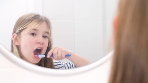 Una bambina si lava i denti con uno spazzolino da denti, un bambino si lava i denti vicino allo specchio in bagno. Concetto di denti sani - Filmati, video