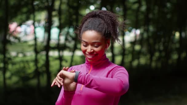 Modern spor aletleri. Genç Afro-Afrikalı Amerikalı kulaklıklı bayan koşucu. Aktivite sonuçlarını akıllı adımlarla kontrol ediyor. Parktaki antrenmandan sonra dinleniyor. - Video, Çekim
