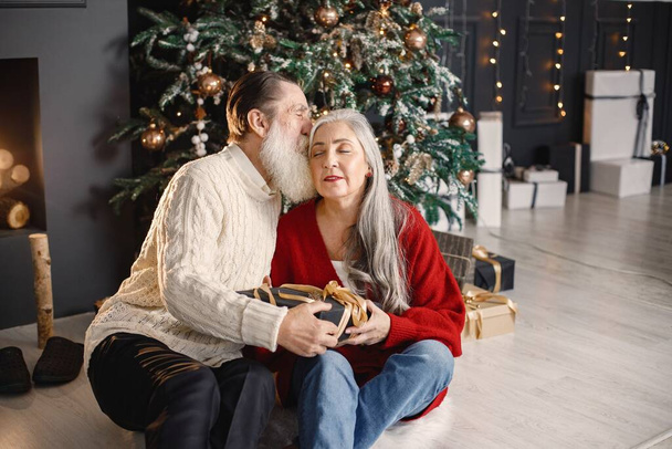 Senior man celebrating christmas with his wife near beautiful christmas tree. Old bearded man sitting on a floor near his wife with grey hair and holding a gifts. Man wearing white sweater and woman - 写真・画像