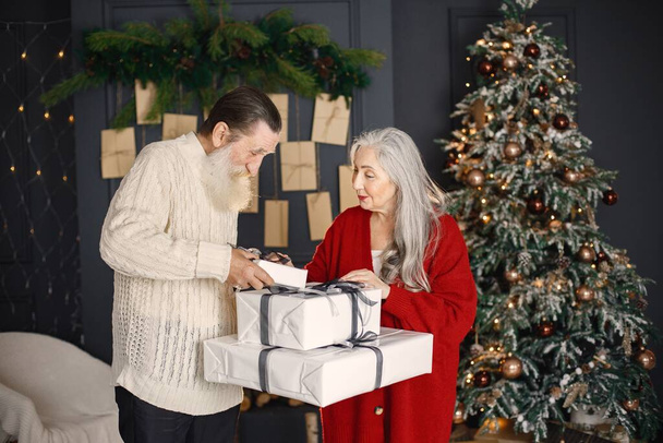 Senior man celebrating christmas with his wife near beautiful christmas tree. Old bearded man standing near his wife with grey hair and holding a gifts. Man wearing white sweater and woman red one. - 写真・画像