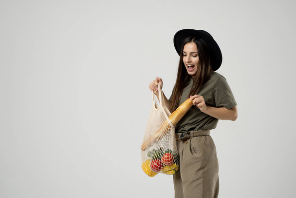 Zero waste concept. A young girl holds on her shoulder a textile mesh eco bag with a groceries. The girl smiles, wearing a beige t-shirt and hat. Refusal of plastic bags - Fotoğraf, Görsel