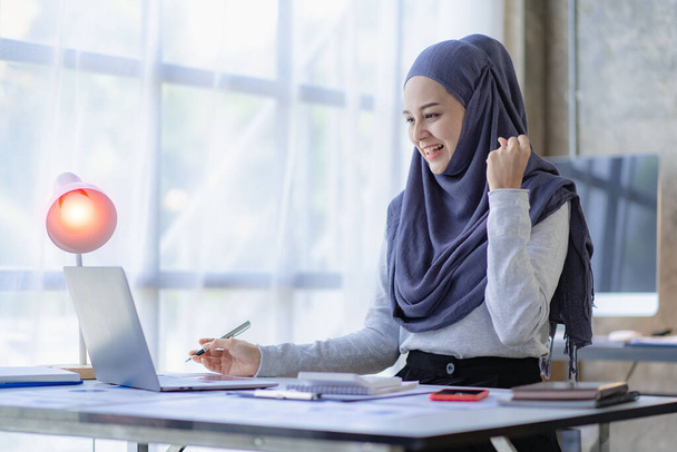 Μια μουσουλμάνα επιχειρηματίας είναι πολύ ενθουσιασμένη που θα κλείσει την πώληση. Αραβίδα γυναίκα χαμογελά και εργάζεται σε φορητό υπολογιστή με οικονομικό διάγραμμα - Φωτογραφία, εικόνα