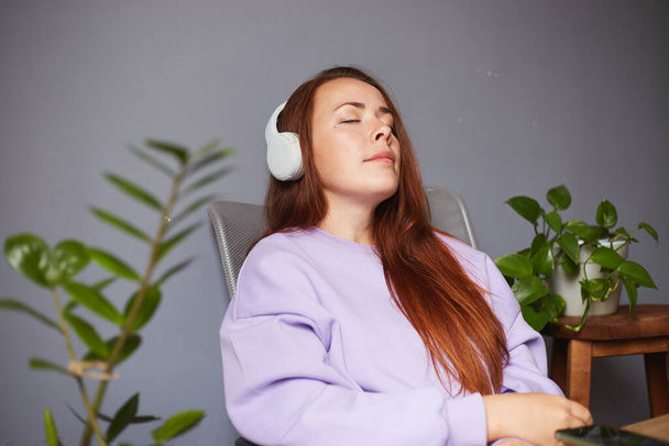 Cheerful caucasian female listening music or podcast and holding smartphone. A lot of green plants around. Red hair woman in violet sweater - Fotografie, Obrázek