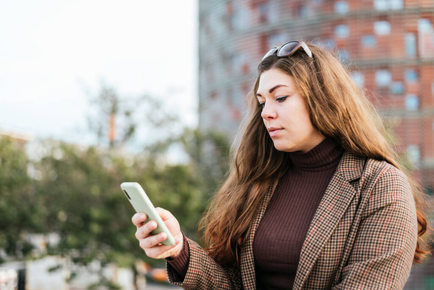 Женщина в верхней одежде с длинными волосами просматривает социальные сети по мобильному телефону, стоя на размытом фоне городской улицы в дневное время - Фото, изображение