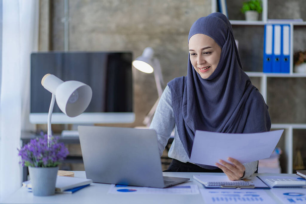 Ασιάτισσα Μουσουλμάνα επιχειρηματίας με μαντίλα χρησιμοποιώντας φορητό υπολογιστή και δουλεύοντας με διαγράμματα, οικονομικά έγγραφα, αναλύοντας λογιστικούς υπολογισμούς στο γραφείο. - Φωτογραφία, εικόνα