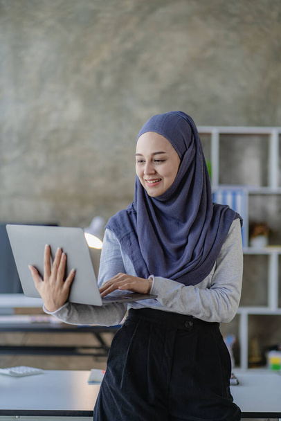 Ασιάτισσα Μουσουλμάνα επιχειρηματίας με μαντίλα χρησιμοποιώντας φορητό υπολογιστή και δουλεύοντας με διαγράμματα, οικονομικά έγγραφα, αναλύοντας λογιστικούς υπολογισμούς στο γραφείο. - Φωτογραφία, εικόνα