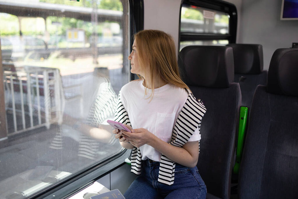 若いかなりブロンドの女性の観光客が電車で窓の近くにスマートフォンを使用して座って旅行する。旅のコンセプトを楽しむ.  - 写真・画像