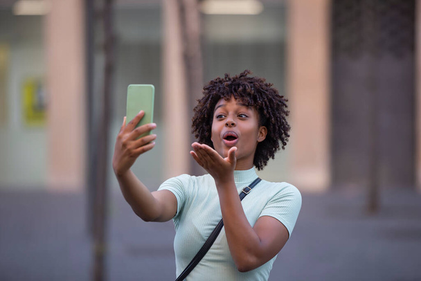 Szczęśliwa kobieta blogger z afro włosów gospodarstwa telefon o rozmowy wideo podczas spaceru w mieście - Zdjęcie, obraz