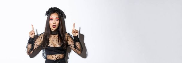 Impressionné fille asiatique en robe de dentelle noire et wreat tourner votre attention sur Halloween promo, faire l'annonce, pointant les doigts vers le haut, montrant la bannière, debout sur fond blanc. - Photo, image
