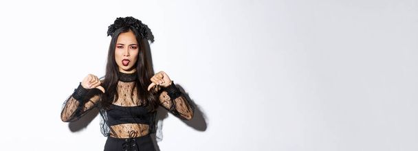 Imagen de mujer asiática decepcionada con disfraz de Halloween de bruja, mostrando pulgares hacia abajo y la lengua pegajosa disgustado, fondo blanco. - Foto, Imagen