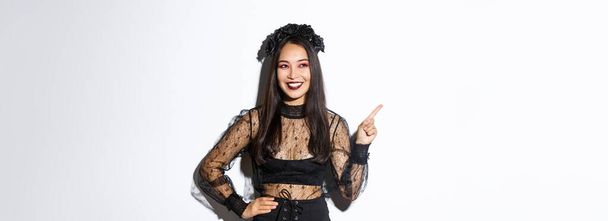 Satisfied smiling asian woman in evil witch or banshee costume celebrating halloween, looking pleased and pointing finger upper left corner, showing your promo banner, white background. - Fotoğraf, Görsel