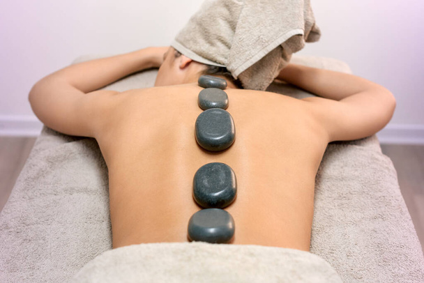 Angle élevé de femelle nue méconnaissable avec des pierres chaudes sur le dos couché sur la table pendant la séance de massage dans le salon de spa - Photo, image