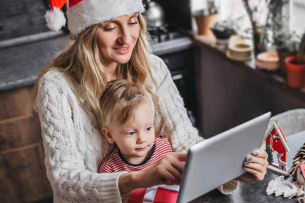 新年の帽子と白いセーターの母親は、彼女の小さな息子にタブレットでゲームを示し、彼と一緒に余暇を過ごします。ママはオンラインストアで彼女の息子のためのおもちゃを注文 - 写真・画像