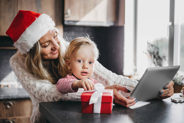 Mother sits with her little son in the kitchen and makes online purchases in the online store. Mom and baby play together on a tablet - Foto, Bild