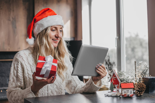 クリスマスビデオ通話。サンタの帽子の若い幸せな女性は、贈り物と赤い箱を保持し、彼女の手にタブレットを保持し、ビデオ会議コールを行います - 写真・画像