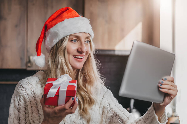 Πρωτοχρονιάτικες βιντεοκλήσεις. Ξανθιά λευκή κοπέλα με εορταστικό χριστουγεννιάτικο καπέλο κρατά ένα τάμπλετ στα χέρια της και ένα κόκκινο κουτί με δώρο, χαμογελάει και κοιτάζει την οθόνη - Φωτογραφία, εικόνα