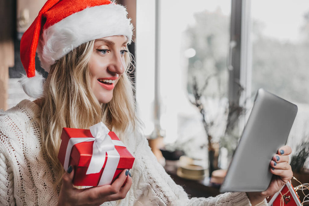 New year video calls concept. Young caucasian blonde woman wearing santa claus festive hat holding a red box with a gift and talking by videoconference remotely during quarantine - Photo, Image