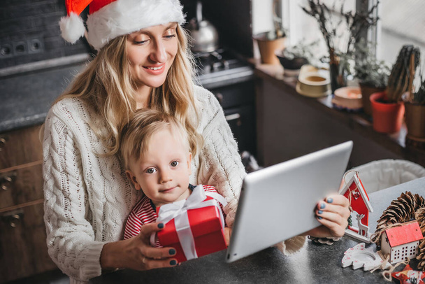 Máma v Santa klobouku sedí s dítětem doma, drží tablet a dělá videokonferenční hovor. Matka a syn obdrželi dárek od svých příbuzných a komunikovali videohovory - Fotografie, Obrázek