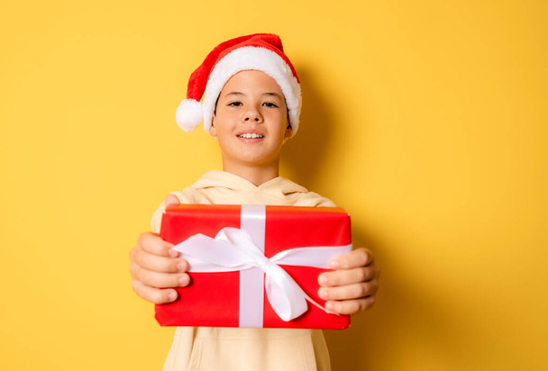 クリスマスの時間-黄色の背景の上にギフトボックスを保持サンタクロース帽子と笑顔の男の子の肖像画を閉じます. - 写真・画像