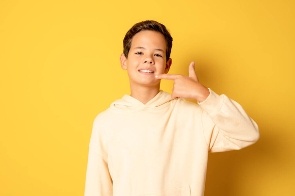 Little boy smiles, pointing finger at mouth standing isolated over yellow background. - Zdjęcie, obraz