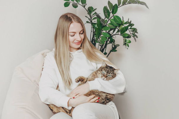 jong blond vrouw houden een tabby donker kat glimlachen en kijken naar hem terwijl zitten op een fauteuil - Foto, afbeelding