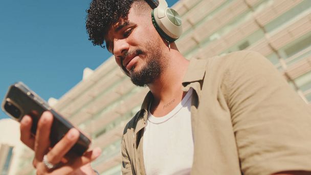 Close up, young male student dressed in an olive color shirt sits outside next to the university, listens to music on headphones, selects tracks on his phone - Photo, image