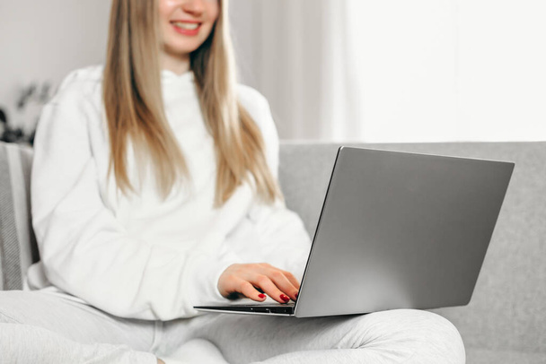 Genç sarışın bir kadın kanepede oturuyor ve laptopunu kullanıyor. Uzaktan eğitim. İnternette çalışıyorum. Serbest Çalışan Kız - Fotoğraf, Görsel