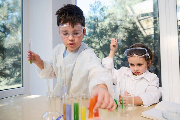 Ragazzi carini, ragazzo e ragazza caucasici in occhiali protettivi e camici, che imparano chimica in laboratorio chimico. Bambini piccoli a lezione. Torniamo a scuola. Studenti in età elementare che fanno esperimenti - Foto, immagini